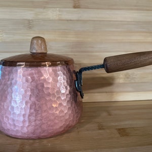 May include: A hammered copper saucepan with a wooden handle and lid. The saucepan has a hammered texture and a light pink hue.