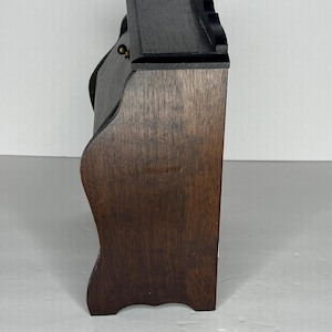 Puede incluir: Una caja de madera marr&oacute;n oscuro con una tapa con bisagras y un peque&ntilde;o pomo de lat&oacute;n. La caja tiene un frente ligeramente curvado y un borde superior festoneado.