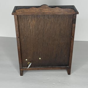 Puede incluir: Una caja de m&uacute;sica de madera con un acabado marr&oacute;n oscuro. La caja tiene una puerta con bisagras y una llave de cuerda en el lateral. La caja est&aacute; sobre una superficie blanca.