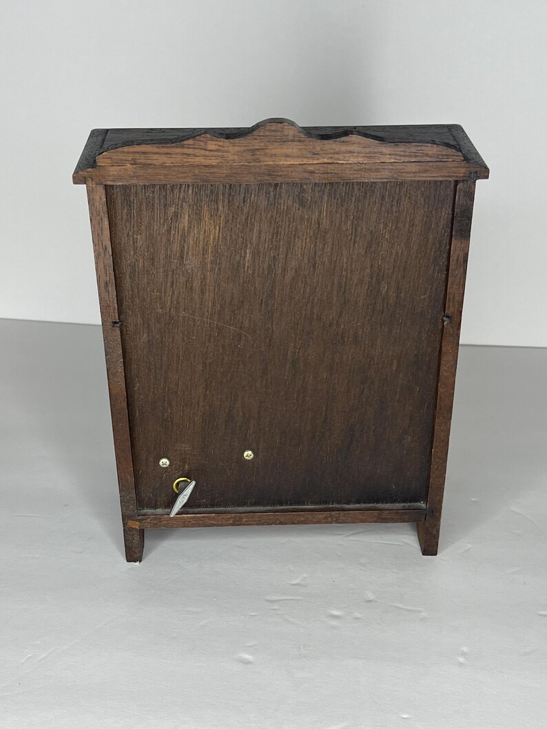 Puede incluir: Una caja de m&uacute;sica de madera con un acabado marr&oacute;n oscuro. La caja tiene una puerta con bisagras y una llave de cuerda en el lateral. La caja est&aacute; sobre una superficie blanca.