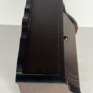 Puede incluir: Una caja de madera marr&oacute;n oscuro con un borde festoneado y una peque&ntilde;a perilla de lat&oacute;n. La caja tiene un borde negro y una incrustaci&oacute;n marr&oacute;n claro.