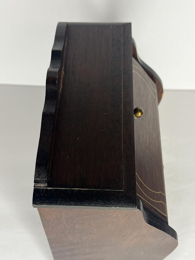 Puede incluir: Una caja de madera marr&oacute;n oscuro con un borde festoneado y una peque&ntilde;a perilla de lat&oacute;n. La caja tiene un borde negro y una incrustaci&oacute;n marr&oacute;n claro.