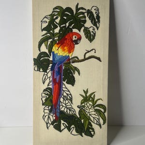 May include: A rectangular wall hanging featuring a detailed embroidery of a vibrant red, yellow, and blue macaw perched on a branch amidst green monstera leaves, all stitched onto a light beige fabric background with a map of Texas and Louisiana.