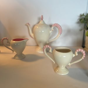May include: A set of four white ceramic teacups with pink floral accents. The set includes a teapot, creamer, sugar bowl, and a cup.
