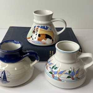 May include: Three ceramic mugs with unique designs. One mug features a sailboat, another has a colorful cat pattern, and the third displays flying ducks. Each mug has a handle and a distinct shape, perfect for coffee or tea.