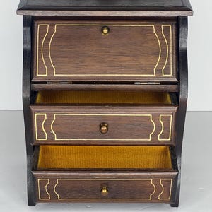 Puede incluir: Una caja de joyer&iacute;a de madera marr&oacute;n oscuro con tapa abatible y tres cajones. Los cajones tienen ribetes dorados y pomos de lat&oacute;n. La caja est&aacute; forrada con terciopelo amarillo.