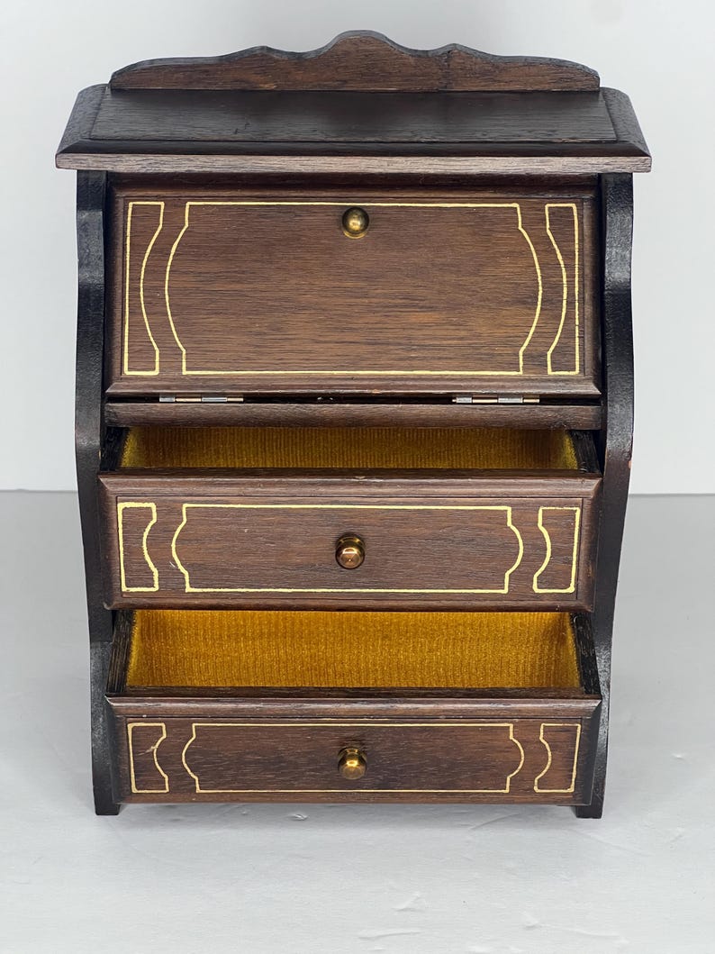 Puede incluir: Una caja de joyer&iacute;a de madera marr&oacute;n oscuro con tapa abatible y tres cajones. Los cajones tienen ribetes dorados y pomos de lat&oacute;n. La caja est&aacute; forrada con terciopelo amarillo.