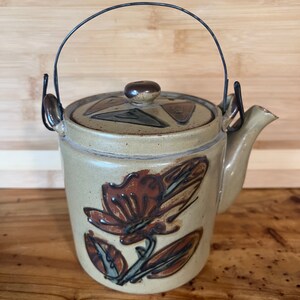 May include: A brown ceramic teapot with a floral design and a wire handle. The teapot has a lid with a geometric pattern.