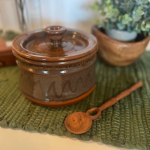 May include: A brown ceramic jar with a lid and a wooden spoon. The jar has a brown glaze with a wavy pattern. The spoon is made of wood and has a round bowl.