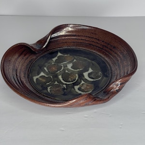 May include: A brown ceramic serving dish with a wavy rim and a glazed interior featuring a pattern of brown and white spots.