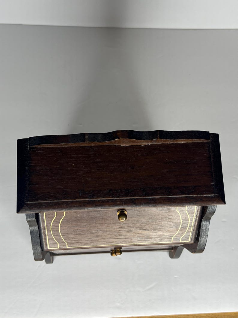 Puede incluir: Una caja de madera marr&oacute;n oscuro con un caj&oacute;n y una tapa con bisagras. La caja tiene un ribete dorado alrededor de los bordes del caj&oacute;n y la tapa.