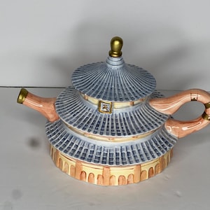 May include: A ceramic teapot in the shape of a pagoda. The teapot features a multi-tiered design with a blue roof, peach-colored accents, and gold-colored details. The handle and spout are peach-colored.