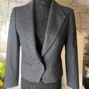 May include: A charcoal gray blazer with a notched lapel, two front pockets, and a single-button closure. The jacket is displayed on a mannequin with a black lace skirt. The background includes a vintage map.