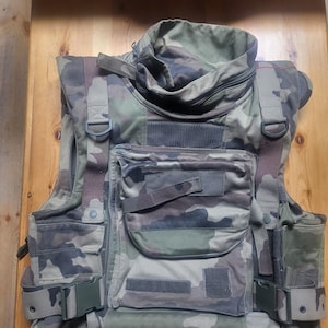 May include: Camouflage tactical vest with multiple pockets, straps, and buckles. The vest has a high collar and is made of a durable, military-style fabric. The colour palette includes shades of green, brown, and tan.