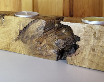 May include: A rustic wooden candle holder with two tealight candle wells. The natural wood features a unique grain pattern and a dark, textured knot. The candles are in silver metal holders, creating a warm, inviting ambiance.