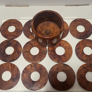 May include: A set of wooden coasters and a matching holder. The coasters are circular with a hole in the centre, and the wood has a rich, dark brown grain. The holder is a cylindrical container, also made of the same wood.