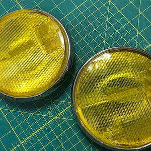 May include: A pair of vintage yellow fog lights with chrome rims. The lights feature a ribbed lens design and a small emblem. The background is a green cutting mat.