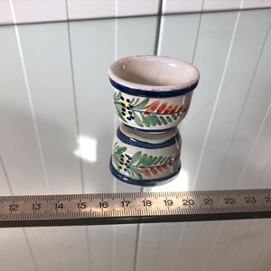 May include: A small, hand-painted ceramic bowl with a white interior and a blue rim. The exterior features a floral design with green leaves, red accents, and blue berries. The bowl is approximately 2 inches in diameter.