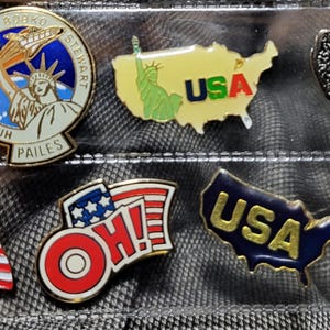 May include: A collection of patriotic enamel pins featuring American flags, the Statue of Liberty, and the USA. The pins are in various shapes and colors, including red, white, blue, and gold. The pins are displayed in a clear plastic case.
