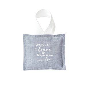 May include: A small, square, blue-gray fabric ornament with white stitching and a white ribbon loop. The text "peace I leave with you" and "john 14:27" are printed in white script on the front.