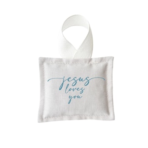 May include: A square, light-colored fabric sachet with a white ribbon loop for hanging. The phrase "jesus loves you" is written in a cursive, light blue font on the front of the sachet. The edges are neatly stitched.