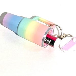 May include: A rainbow-colored travel mug with a built-in lip balm holder. The mug has a silver lid and a keychain attached. The lip balm is pink and says "Slick" on the label.