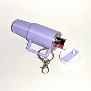 May include: A purple keychain with a small container that holds a lip balm. The lip balm is red and white and says "Tick Protectant Cherry". The keychain has a silver ring and a silver clip.
