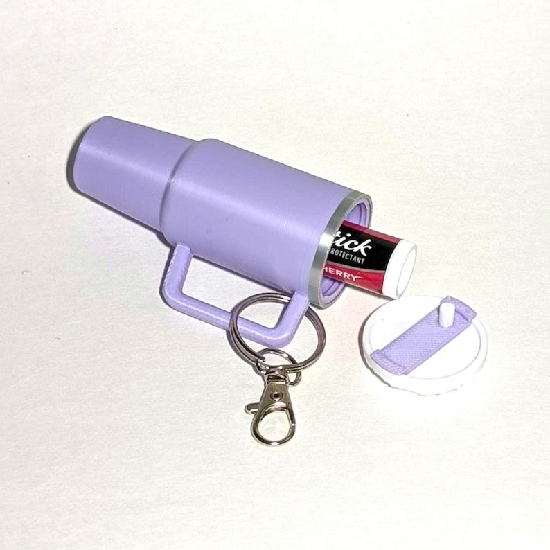 May include: A purple keychain with a small container that holds a lip balm. The lip balm is red and white and says "Tick Protectant Cherry". The keychain has a silver ring and a silver clip.