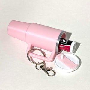 May include: A pink plastic keychain mug with a removable lid. The mug has a silver metal band around the top and a silver metal keychain attached. The lid is open and a small tube of lip balm is inside. The lip balm is labeled "Tick Protectant" and "Berry".