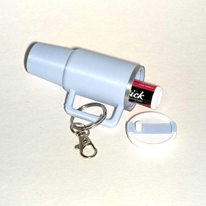 May include: A light blue keychain with a silver ring and a clip. The keychain has a compartment that holds a lip balm tube. The lip balm tube is labeled "Tick" and "Protectant".