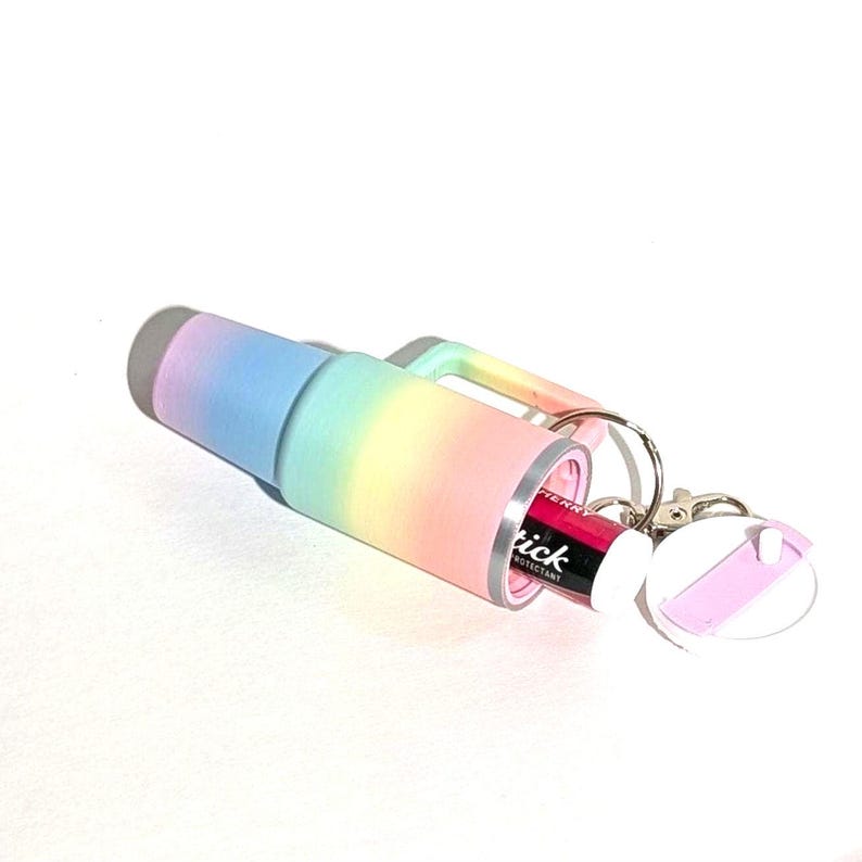 May include: A rainbow-colored travel mug with a built-in lip balm holder. The mug has a silver lid and a keychain attached. The lip balm is pink and says "Slick" on the label.