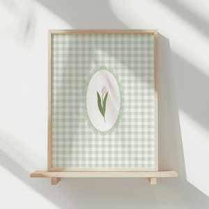 Könnte beinhalten: Ein gerahmter Druck mit hellgrünem Vichy-Muster im Hintergrund. Ein ovales Design in der Mitte zeigt eine rosa Tulpe mit grünen Blättern, umrahmt von einem spitzenartigen Rand. Der Holzrahmen ruht auf einem Holzregal.