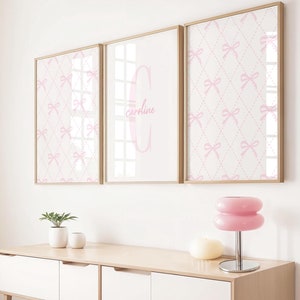 Könnte beinhalten: Drei gerahmte Drucke mit einem rosa-weißen Schleifenmuster. Der mittlere Druck zeigt den Namen "Caroline" in rosa Schrift. Die Drucke hängen an einer weißen Wand über einer Kommode aus Holz mit zwei Schubladen.