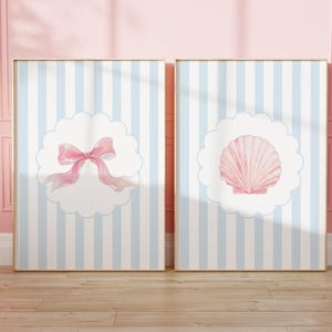 May include: Two framed art prints. One features a pink bow, the other a pink seashell, both set against a blue and white striped background. The prints are in gold frames, displayed on a wooden floor against a pink wall.