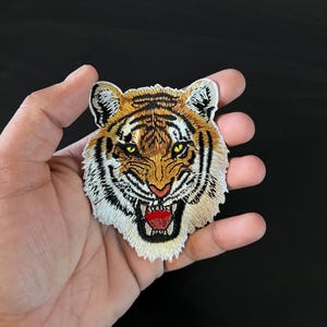 May include: A close-up of an embroidered tiger face patch. The tiger has orange and black stripes, white fur, and a red open mouth.