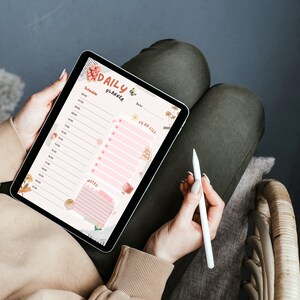 May include: A digital tablet displaying a "Daily Planner" layout with a schedule, to-do list, and notes section. The planner has a pink and beige color scheme with floral illustrations. A white stylus rests on the right.