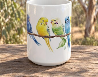 Peut inclure: Un mug en céramique blanche orné d'une illustration à l'aquarelle de plusieurs perruches ondulées bleues, jaunes et vertes perchées sur une branche brune qui fait le tour de la surface extérieure du récipient cylindrique.