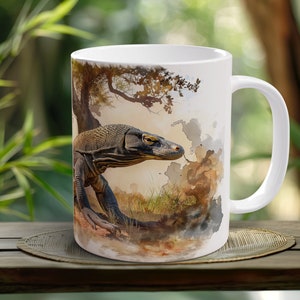 Puede incluir: Taza de cerámica blanca con una ilustración de acuarela de un dragón de Komodo en un entorno de jungla. El dragón es marrón y negro con una cola larga y garras afiladas.