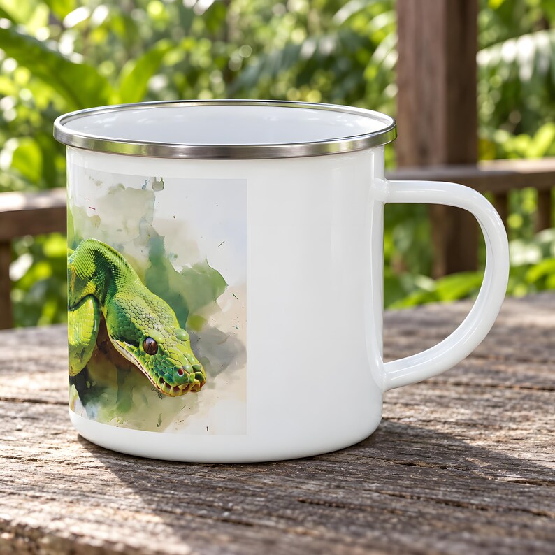 May include: A white enamel mug featuring a watercolor-style illustration of a green snake on the side. The mug has a smooth white finish, a silver-colored metal rim, and a curved handle on the right side, presenting a clean and functional design.