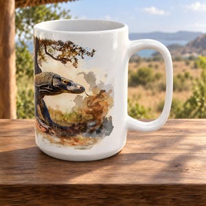 May include: A white ceramic mug featuring a detailed watercolour-style illustration of a Komodo dragon. The lizard is depicted in shades of brown, grey, and black, set against a soft, abstract background of earthy tones and hints of green foliage.
