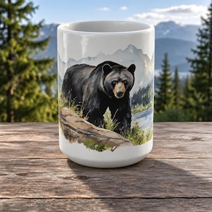 Könnte beinhalten: Ein weißer Keramikbecher mit einer Aquarell-Illustration eines Schwarzbären, der auf einem Felsvorsprung an einem Fluss steht, vor einer Kulisse aus fernen Bergen und immergrünen Bäumen in sanften, gedämpften Farbtönen.