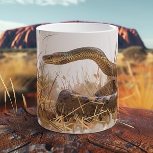 Puede incluir: Taza de cerámica blanca con una ilustración detallada de una serpiente en un campo de hierba, con un fondo borroso de una formación rocosa roja y un cielo azul. La taza está colocada sobre una superficie de madera, creando un diseño temático de la naturaleza.