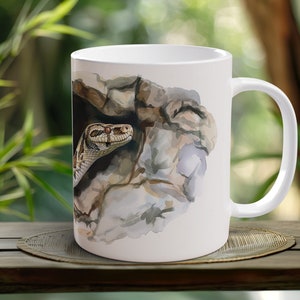 Könnte beinhalten: Weiße Keramiktasse mit einer Aquarell-Illustration einer Schlange, die aus einer Felsspalte schaut.