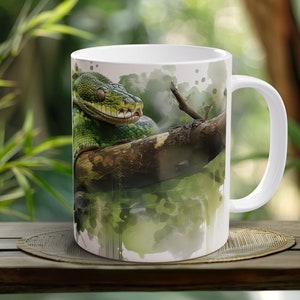 May include: White ceramic mug with a detailed illustration of a green snake on a tree branch. The snake's scales and the branch's texture are visible. The background has a watercolor effect with green and white hues.
