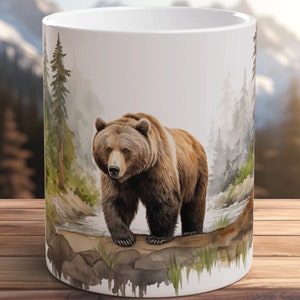 Puede incluir: Una taza de cerámica blanca con una ilustración de acuarela de un oso marrón de pie sobre rocas en un entorno de bosque. El oso está mirando directamente al espectador.