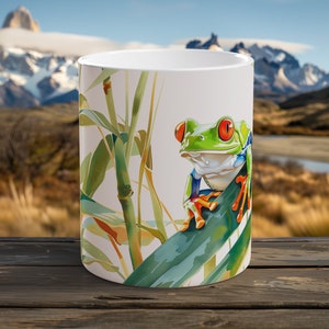 May include: White ceramic mug featuring a vibrant watercolor illustration of a red-eyed tree frog perched on a leaf. The frog has bright green skin, red eyes, and orange feet. Bamboo stalks and leaves are also depicted on the mug.