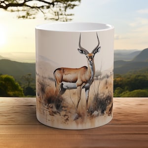 Könnte beinhalten: Weiße Keramiktasse mit einer braunen und weißen Illustration eines Antilopen, das in einem grasbewachsenen Feld steht. Der Hintergrund ist ein verschwommenes Bild einer Bergkette.