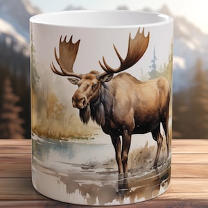 May include: White ceramic mug with a watercolor illustration of a brown moose standing in a lake. The moose has large antlers and is surrounded by green foliage.