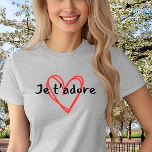 May include: A gray t-shirt with a red heart and the text "Je t'adore" printed on the front. The shirt is being worn by a person in a park setting.