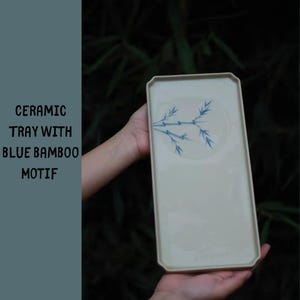 May include: A rectangular ceramic tray with a light beige surface and a blue bamboo motif. The tray has a slightly raised edge and is held by a person. The text on the left says "CERAMIC TRAY WITH BLUE BAMBOO MOTIF".